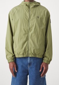 Ljusgrön vindjacka med framzipper, elastiska ärmslut och huva. Har logotyp på bröstet och en avslappnad passform.