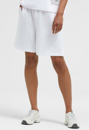 Person, die weiße knielange Shorts, weiße Sneaker und einen Teil eines weißen Oberteils trägt, steht vor einem schlichten hellen Hintergrund.