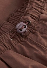 Shorts en tissu marron avec taille élastique, fermeture à cordon de serrage, et texture froncée sur le dessus, comprenant un ajusteur à bouton-pression et des œillets en métal.