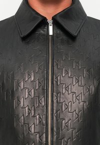 Juodas odinis švarkas su išdegtomis ornamentais, turintis smailėjančią apykaklę ir priekinį užtrauktuką su metaliniais akcentais.