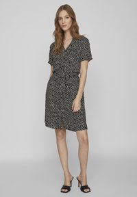 Donna in piedi con capelli lunghi, vestita con un abito a maniche corte avvolgente nero con pois bianchi e mules nere con tacco, contro uno sfondo grigio uniforme.