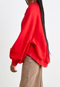 Red oversized sweatshirt with ribbed hem and sleeves; features a relaxed fit and raw edge detailing. Paired with beige wide-leg pants.