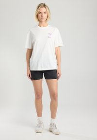 T-shirt en coton blanc avec un design graphique violet, assorti à un short en denim foncé. Modèle portant des baskets blanches et des chaussettes.