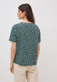 Camiseta de manga corta en verde oscuro con un patrón de hojas, hecha de tela suave. Presenta un cuello redondo y un corte holgado. Se muestra la vista trasera.