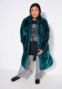 Cappotto in peluche color teal con colletto, due tasche frontali e bottoni; indossato sopra una maglietta nera con grafica e pantaloni argentati lucidi.