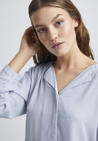 Blusa azzurro chiaro con una texture morbida, caratterizzata da un colletto a scialle e polsini con bottone. Design semplice che si concentra su eleganza e comfort.