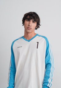T-shirt de sport à manches longues en blanc avec des manches bleu clair, présentant un col rond, trois rayures sur les manches et un "1" imprimé en marron foncé.