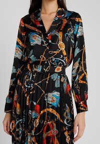 Černé šaty s dlouhým rukávem s barevným vzorem řetězů, pásků a doplňků v červených, modrých a zlatých tónech; plisovaný design.