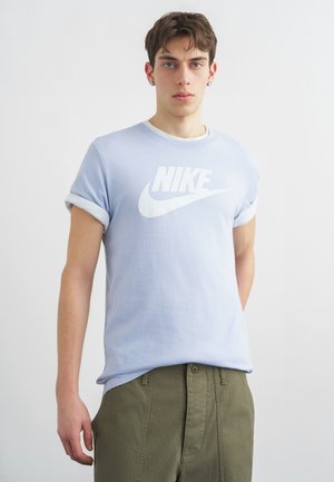 Νεαρός άνδρας με κοντά καστανά μαλλιά, φορώντας ανοιχτό μπλε μπλουζάκι Nike και χακί παντελόνι, στέκεται μπροστά σε λευκό φόντο.