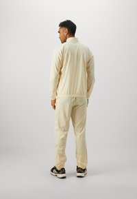 Beige tracksuit with a zip-up jacket and elastic waistband pants. Features elastic cuffs, side pockets, and black accent details.