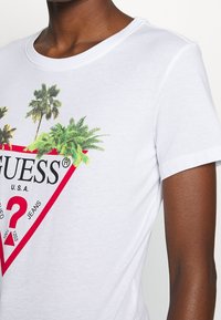 Bílý bavlněný tričko s červenou trojúhelníkovou grafikou se palmami, textem "GUESS" a černými a červenými akcenty. Krátké rukávy.