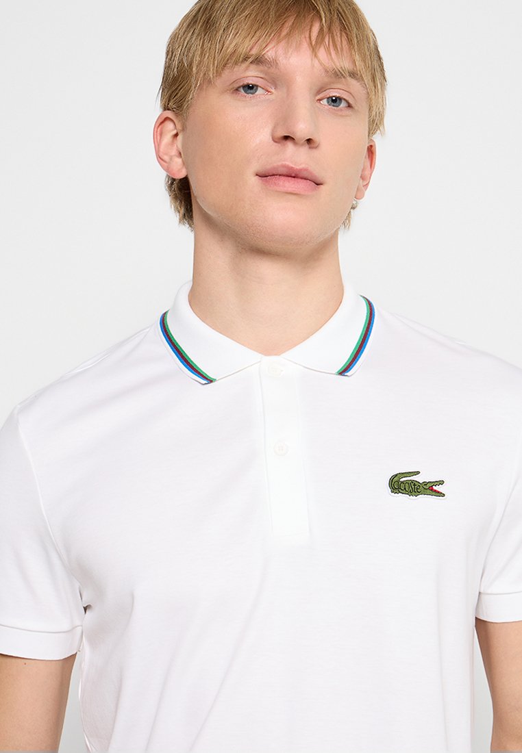 Un jeune homme portant un polo blanc avec un col rayé multicolore et un logo de crocodile vert sur la poitrine gauche.