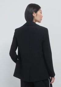 Donna con capelli neri corti, che indossa un blazer nero e pantaloni abbinati con strisce bianche, in piedi contro uno sfondo grigio chiaro.