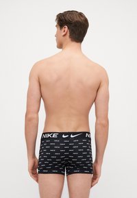 Schwarze Baumwollboxershorts mit sich wiederholendem weißen Nike-Logo-Muster und elastischem Bund, getragen von einem männlichen Modell, das mit dem Rücken zur Kamera steht.