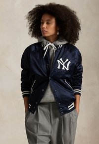 Göndör hajú nő, aki egy tengerészkék szatén New York Yankees dzsekit visel szürke kapucnis pulóver és szürke redős nadrág felett, kezét a zsebében tartva.