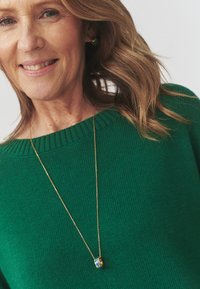 Grøn strikket sweater med ribbede kanter; guld kæderekæde med en lille rund vedhæng med blå, grønne og gule mønstre.