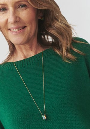 Green knitted sweater with ribbed neckline; gold chain necklace featuring a small rounded pendant with blue, green, and yellow patterns.