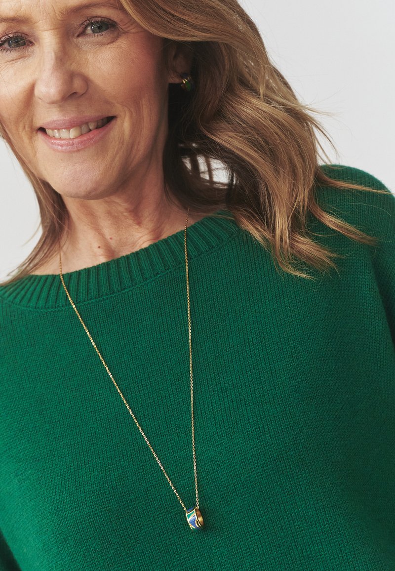 Grøn strikket sweater med ribbede kanter; guld kæderekæde med en lille rund vedhæng med blå, grønne og gule mønstre.