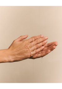 Un gel translucide est versé entre deux mains ; des tons de peau chauds et des textures lisses sont visibles. Du gel goutte le long des doigts. L'arrière-plan est neutre.