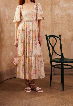 Robe longue fleurie avec manches bouffantes, dans des tons pastel de rose et jaune. Présente un corsage froncé et des volants à étages. Portée avec des sandales blanches.