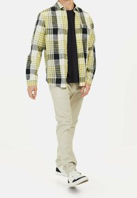 Camicia a quadri gialla, nera e bianca; abbinata a una camicia nera, pantaloni beige chiaro e sneakers bianche con dettagli neri.