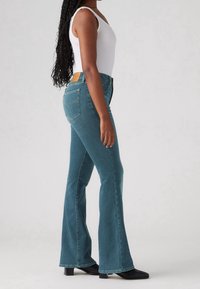 Jeans acampanados en denim azul verdoso con una forma ajustada. Presentan una etiqueta de cuero color beige claro en la parte trasera de la cinturilla y en los bolsillos traseros.