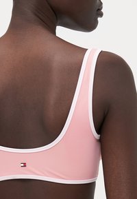 Rosa sports-bh med hvit kant, racerback-design og en liten logo-detalj på baksiden. Jevn tekstur, tettsittende form.