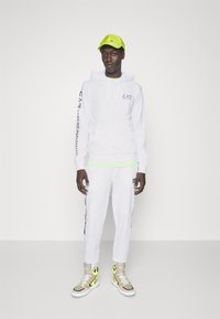 Felpa bianca con logo EA7, abbinata a pantaloni jogger bianchi con accenti laterali. Cappello verde brillante e sneakers alte in beige con dettagli neon.