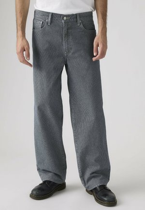 Man wearing loose-fitting gray denim jeans, black shoes, and a white shirt against a neutral background.