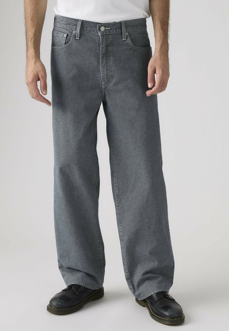 Mand iført løstsiddende grå denimjeans, sorte sko og en hvid skjorte mod en neutral baggrund.