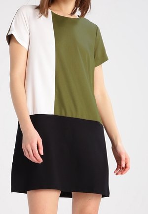 Kobieta w sukience z krótkim rękawem w kolorach białym, oliwkowozielonym i czarnym, stojąca na jasnym, jednolitym tle.