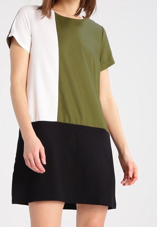 Femme portant une robe à manches courtes en blocs de couleurs blanc, vert olive et noir, debout devant un fond clair uni.