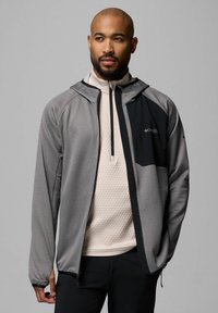 Męska kurtka z kapturem, szary materiał z fakturowanym wzorem, czarne akcenty na klatce piersiowej oraz pełny zamek z przodu, zestawiona z beżową bluzką.