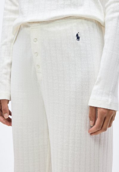 Άσπρο παντελόνι με πλεκτή ραφή και κουμπιά στην εμπρός πλευρά, με μαύρο κεντημένο λογότυπο παικτή πόλο, φορεμένο με ταιριαστό μακρυμάνικο τοπ, με το χέρι χαλαρά στο πλάι.