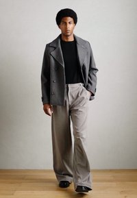 Manteau en laine gris, à double boutonnage, sur un t-shirt noir. Pantalons plissés gris clair, à jambes larges, avec des chaussures noires.