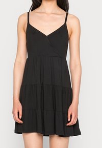 Kobieta ubrana w bezrękawową czarną mini sukienkę z falbanami, z cienkimi ramiączkami i dekoltem w kształcie litery V.