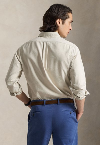 Ανοιχτό μπεζ πουκάμισο με ρυτιδιασμένα μανίκια, λεπτομέρεια πιέτας στο πίσω μέρος, συνδυασμένο με μπλε παντελόνια που διαθέτουν πλαϊνές και πίσω τσέπες και καφέ ζώνη.