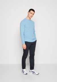 Un giovane uomo è in piedi contro un muro bianco indossando un maglione azzurro chiaro, pantaloni scuri e sneakers bianche con dettagli blu, con le mani in tasca.