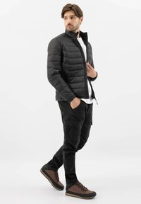 Uomo che indossa una giacca nera trapuntata, una camicia bianca, pantaloni cargo neri e scarpe da trekking marroni, in piedi con le mani in tasca, che guarda di lato.