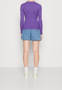 Persona in piedi di spalle con una maglietta a maniche lunghe a coste viola, pantaloni shorts di denim blu, calze bianche e sneakers beige su un pavimento bianco.
