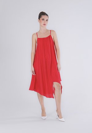 Jonge vrouw staand, gekleed in een losse rode mouwloze jurk met dunne bandjes en witte puntige hoge hakken, tegen een effen lichte achtergrond.