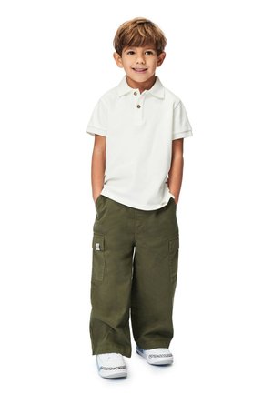 Hvid polo shirt med to knapper, olivengrøn cargo-bukser med lommer, og hvide sneakers med mønstrede detaljer. Barnet står med hænderne i lommerne.