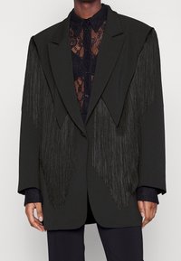 Blazer noir oversize avec détails à franges sur les épaules, forme structurée et tissu lisse, porté sur une chemise en dentelle noire.