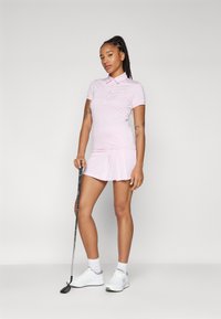 Súprava polo trička a sukne v svetlo ružovej farbe, vyrobená z textúrovaného materiálu. S golierom, gombíkmi a skladanou sukňou. Biele golfové topánky a golfová palica sú súčasťou.