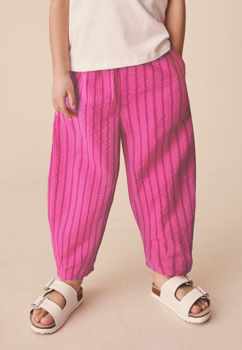Niño con pantalones anchos de rayas rosas, sandalias blancas con hebillas y una camisa blanca, de pie sobre un suelo de color claro.