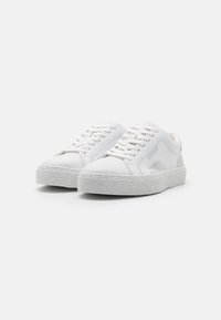 Par de zapatillas bajas blancas con suelas de goma texturizada y cordones blancos sobre un fondo liso claro.