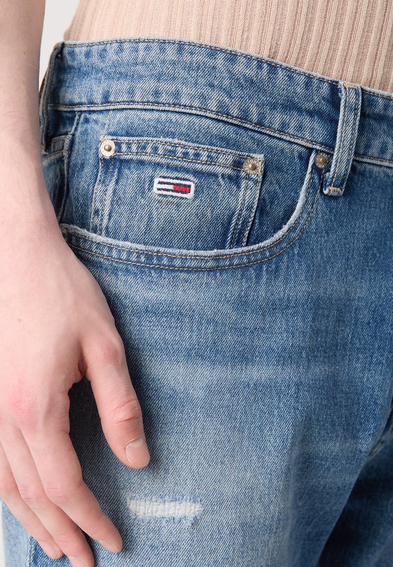 Hellblaue Denim-Jeans mit einem beschädigten Patch, einem kleinen Logo auf der vorderen Tasche und standardmäßigen Metallteilen.