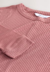 Ružičasta majica s rebrastim uzorkom i dugim rukavima, ovratnika s okruglim izrezom i teksturiranog materijala. Ima vidljive šavove i malu etiketu na ovratniku.
