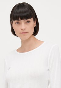 Fehér, hosszú ujjú felső, finom bordázott textúrával és lekerekített nyakkal. Az anyagon apró minták találhatók, amelyek elegáns megjelenést kölcsönöznek.