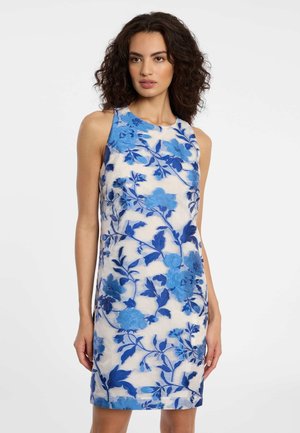 Frau in ärmellosem weißen Kleid mit blauem Blumenmuster, stehend vor einem schlichten hellen Hintergrund.